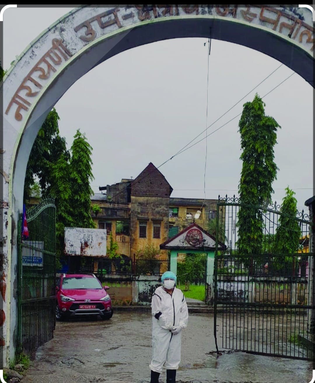 A doctor in full personal protective equipment — coverall, mask, cap, gloves and boots — standing alone inside the gateway arch of Narayani Hospital, Birgunj, on a wet monsoon day.